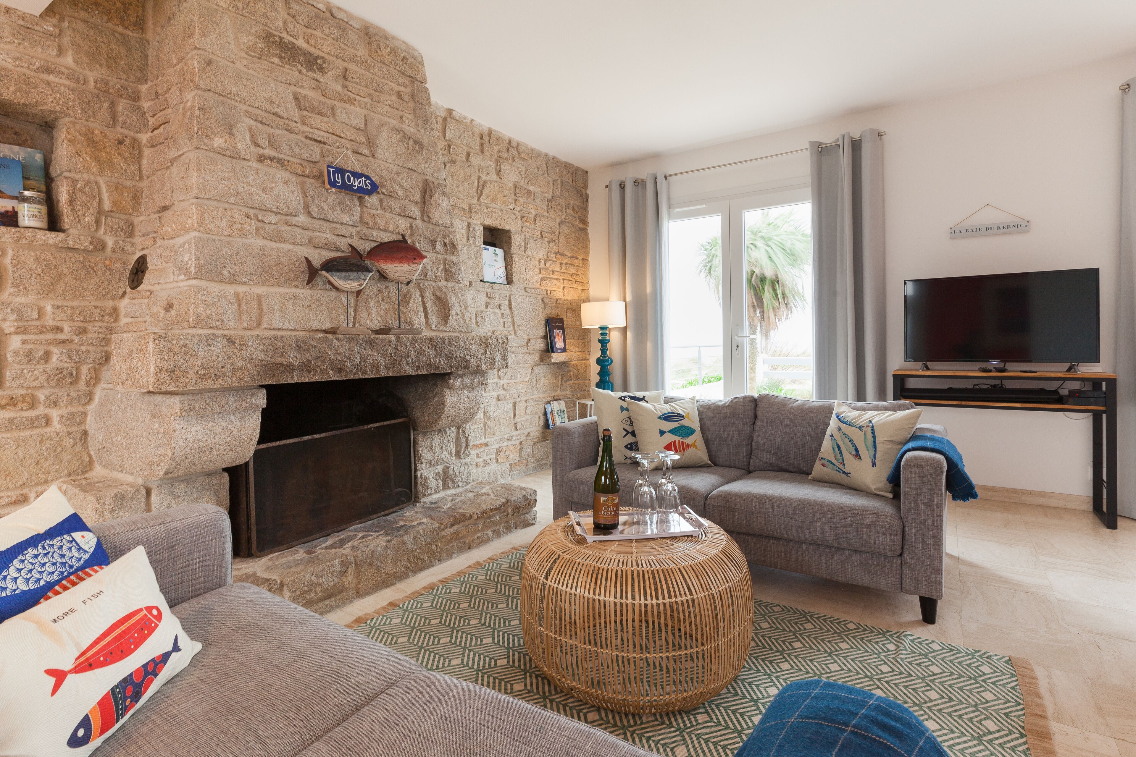 A living room with a stone fireplace, a grey sofa, a round wicker coffee table, and a flat-screen TV.