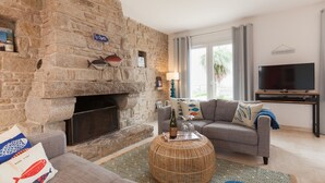 Ein Wohnzimmer mit Kamin aus Stein, einem grauen Sofa, einem runden Korbtisch und einem Flachbildfernseher.