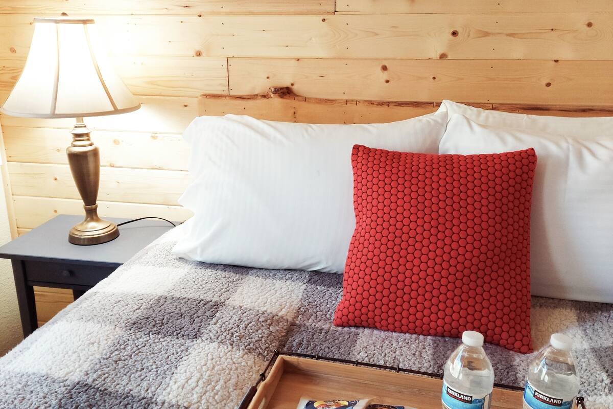 A wooden bed with white and red pillows, a bedside lamp, a tray with a magazine and water bottles.