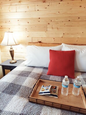 A wooden bed with white and red pillows, a bedside lamp, a tray with a magazine and water bottles.