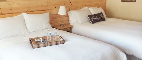 A hotel room with two beds, wooden headboard, white bedding, a bedside table with a lamp, and a tray with water bottles and snacks.