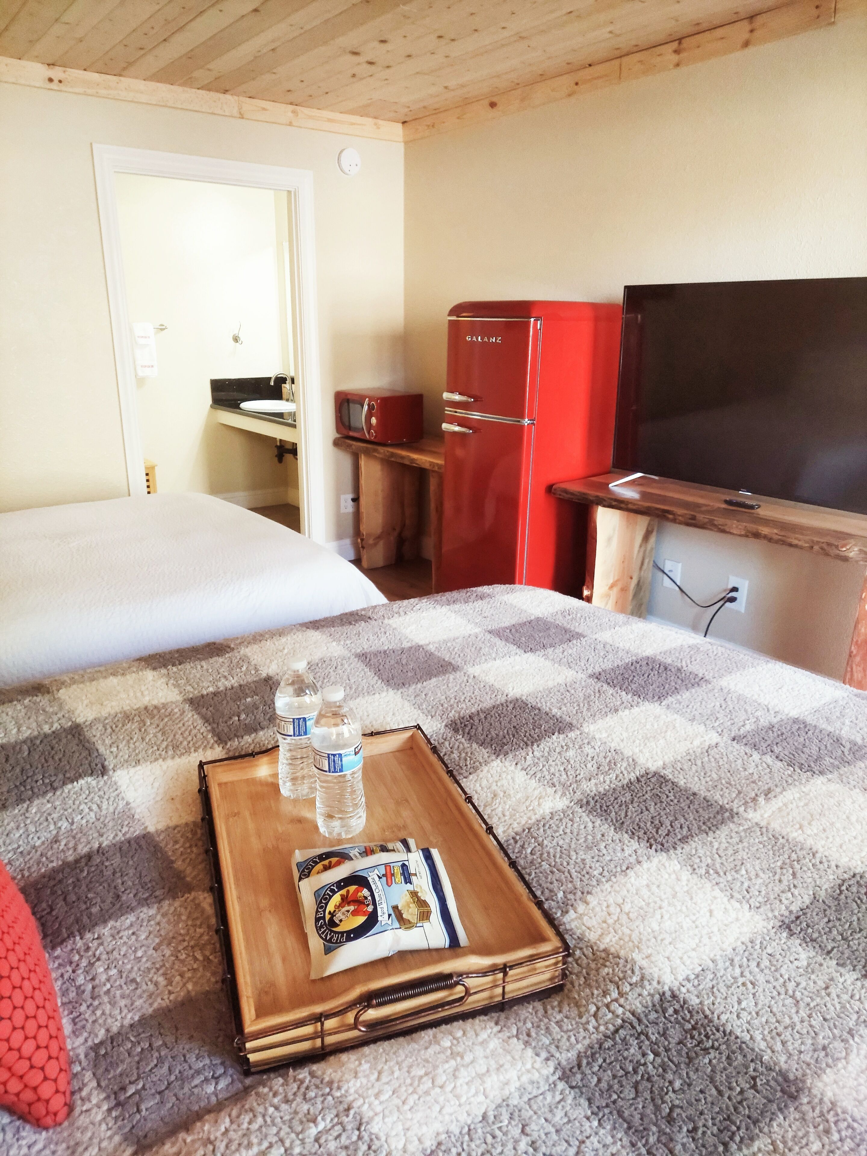 A hotel room with a bed, a wooden bedside table with a tray of bottled water and a packet of biscuits, a red refrigerator, a television, and a wooden cabinet.