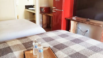 A hotel room with a bed, a wooden bedside table with a tray of bottled water and a packet of biscuits, a red refrigerator, a television, and a wooden cabinet.