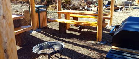 Une structure en bois formant une pergola, avec une table de pique-nique et un espace barbecue, le tout intégré dans un environnement naturel.
