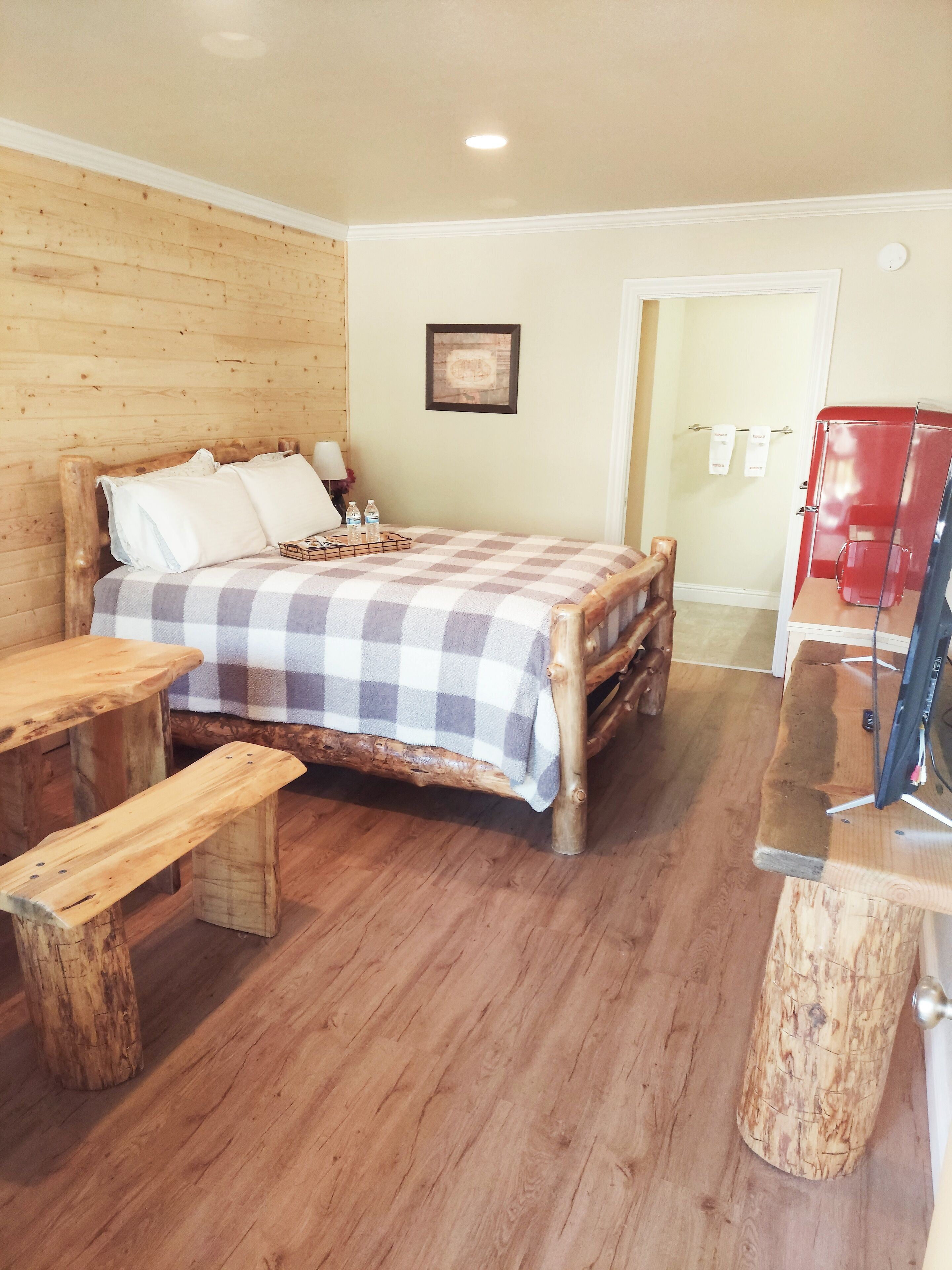 A wooden bed with a checkered blanket, a wooden bench, and a red appliance in a room with wooden flooring and walls.