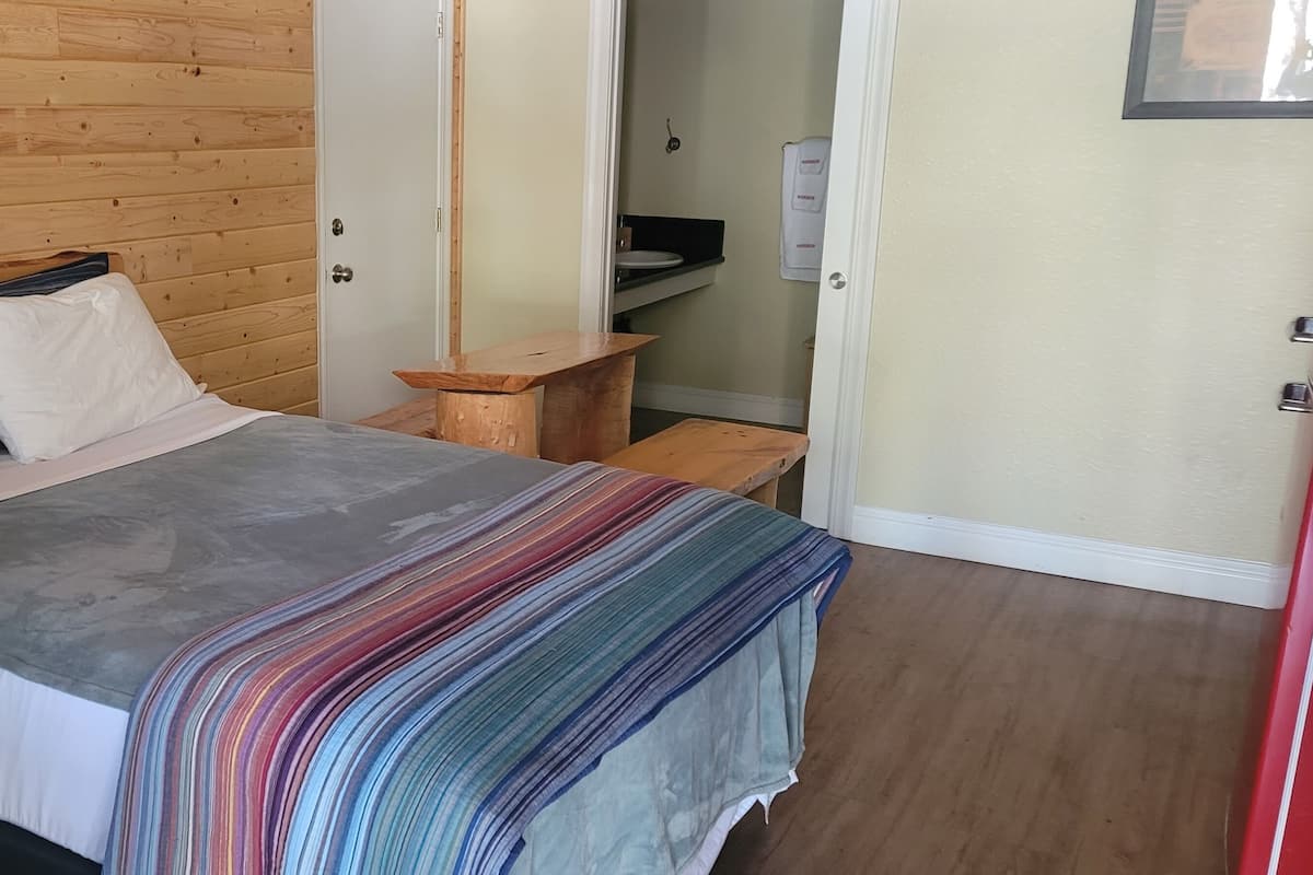 A bedroom with a wooden headboard, a bed with white and gray bedding, a colorful striped blanket, and a small wooden nightstand.