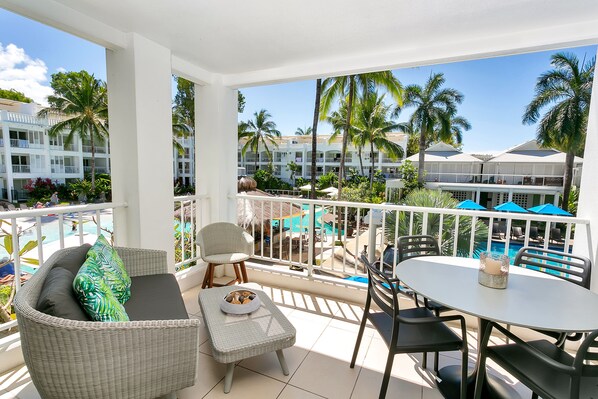 A balcony with wicker furniture, a dining table, and a view of a pool area with palm trees and hotel buildings. - Luxurious 2.5 Bed APT with Fab Beach Club Resort Views (Cairns)