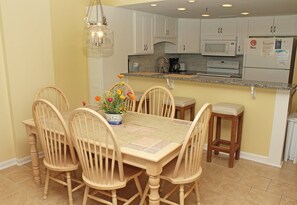 A dining area with a wooden table and chairs, a potted plant, and a kitchen with white cabinets and appliances. - 3rd Floor Oceanfront Condo, Jacuzzi Tub, Outdoor Pool | Crescent Tower II - 306 (North Myrtle Beach)