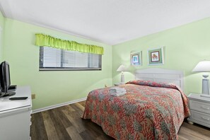 A bedroom with a bed, a desk, a TV, a window with blinds, and two framed pictures on the wall. - 3rd Floor Oceanfront Condo, Jacuzzi Tub, Outdoor Pool | Crescent Tower II - 306 (North Myrtle Beach)