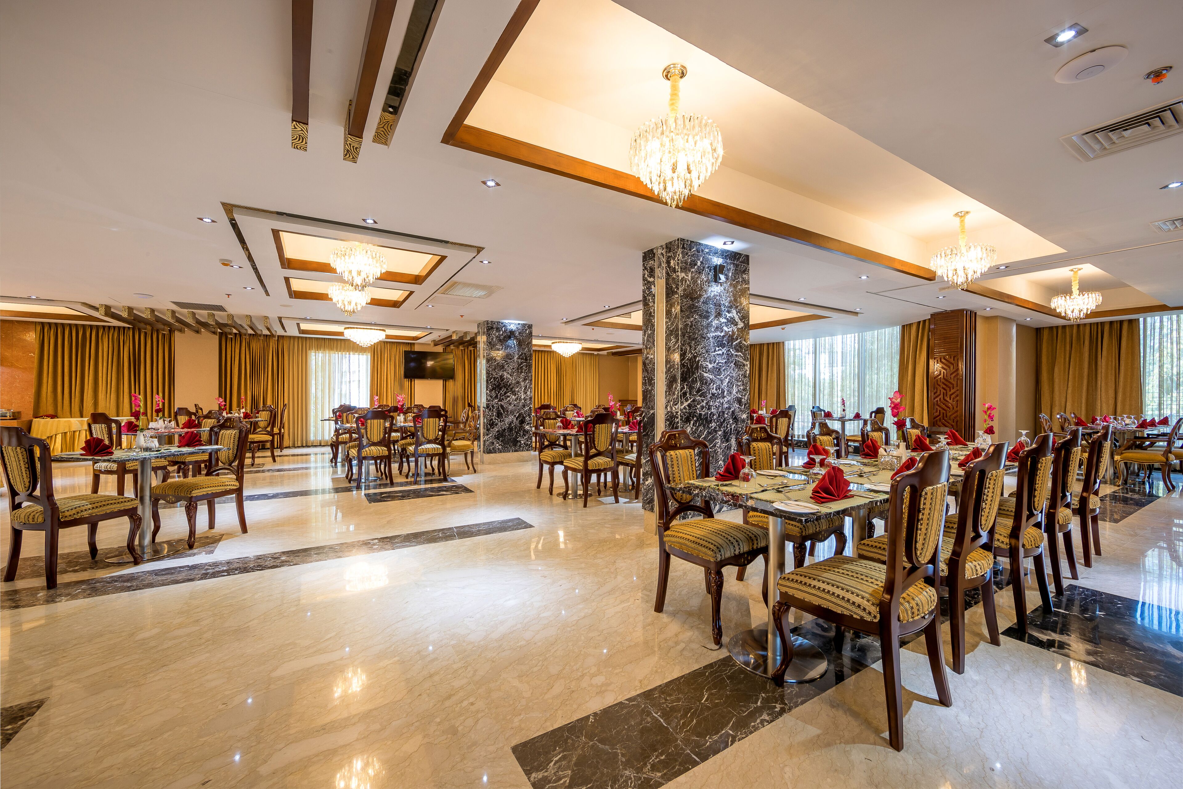 A spacious banquet hall with multiple dining tables and chairs, marble flooring, and chandeliers.