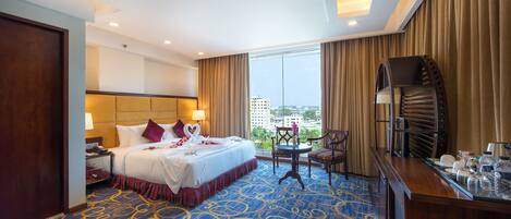 A hotel room with a large bed, a desk with a chair, a view of the city, and a balcony.
