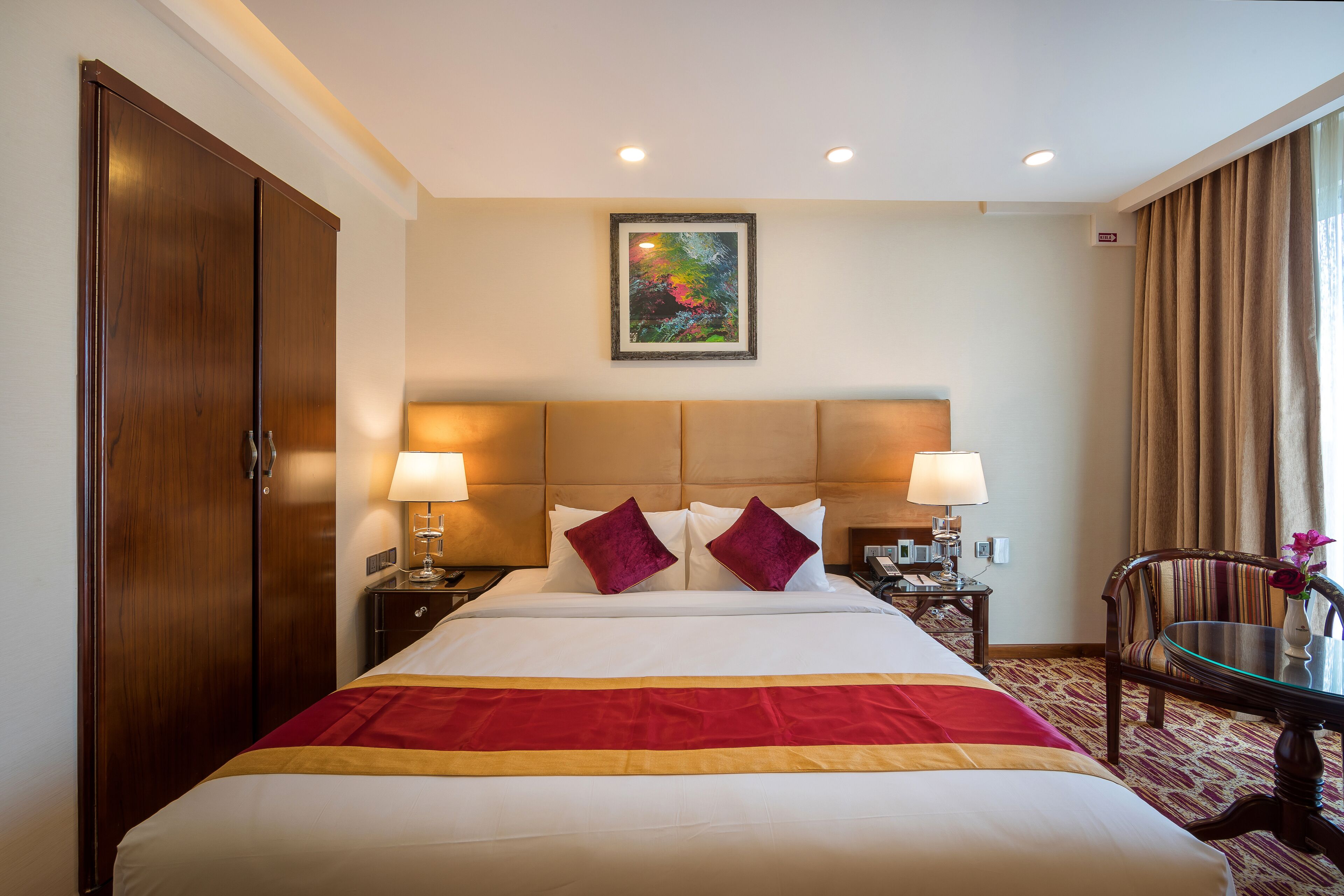 Une chambre d’hôtel avec un grand lit, deux lampes de chevet, une armoire en bois, une œuvre d’art encadrée au mur et une fenêtre avec des rideaux.