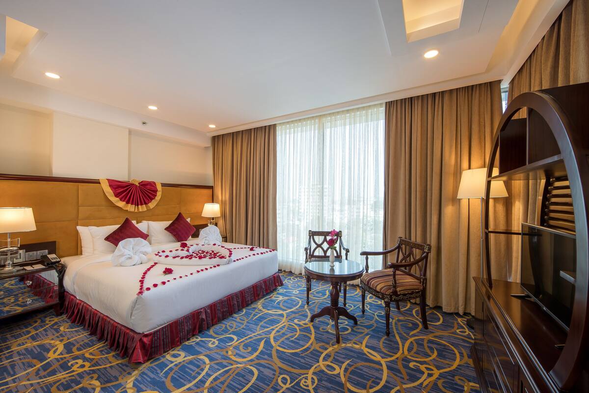 Ein Hotelzimmer mit einem großen Bett, einem Schreibtisch mit Lampe, einem Sessel, einem Fernseher und einem Fenster mit Vorhängen.