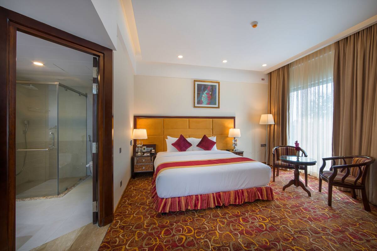 Ein Hotelzimmer mit einem großen Bett, zwei Nachttischlampen, einer Kopfteil, einer Duschkabine, einem kleinen Tisch mit Stuhl und einem großen Fenster mit Vorhängen.