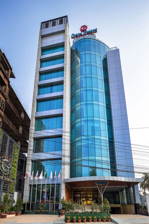 Een modern hotel met een gebogen glazen gevel, een opvallend 'Grand Palace' bord en een rij vlaggen bij de ingang.