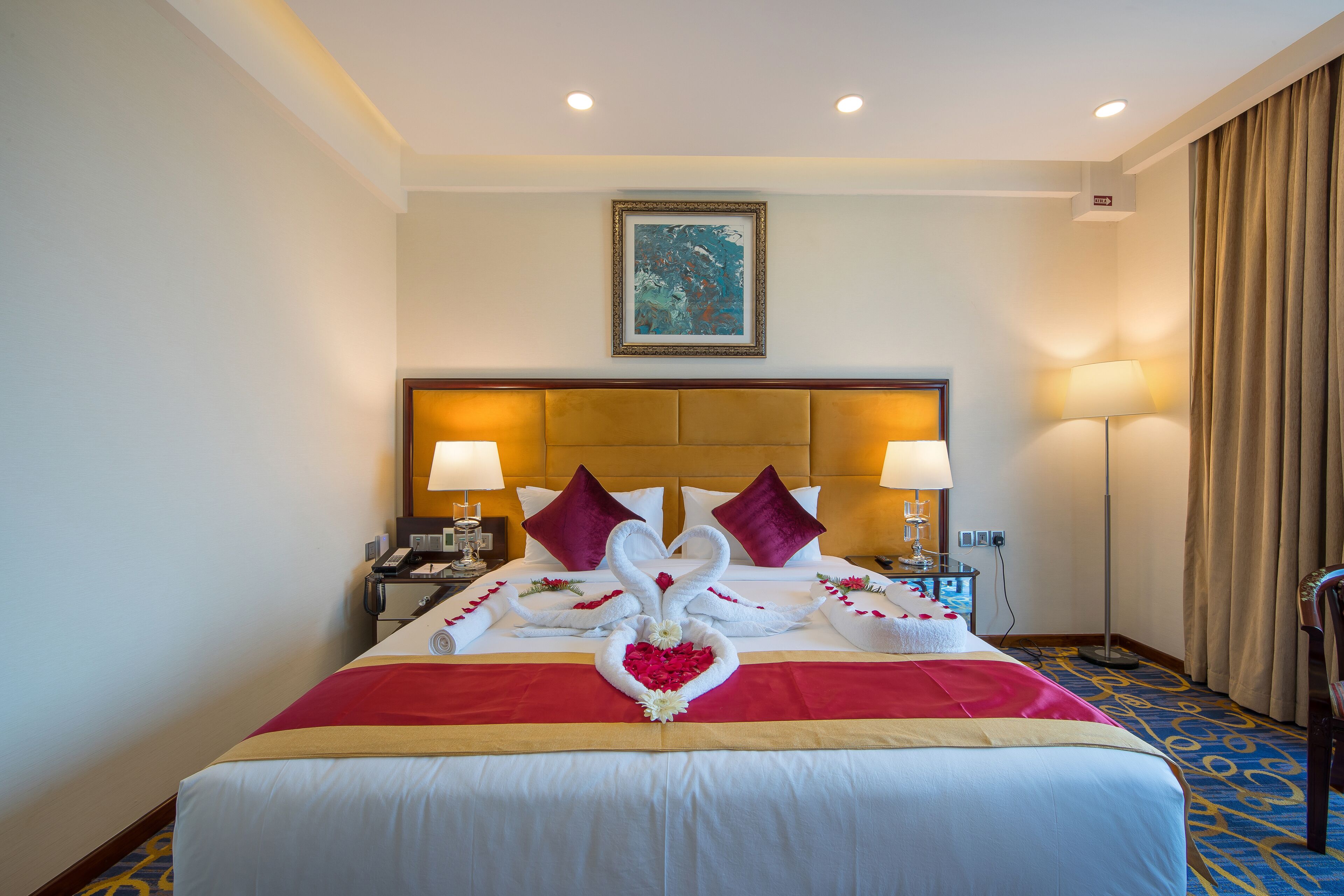 A hotel room with a large bed, two bedside lamps, a headboard, a framed picture on the wall, and a red and white bedspread.