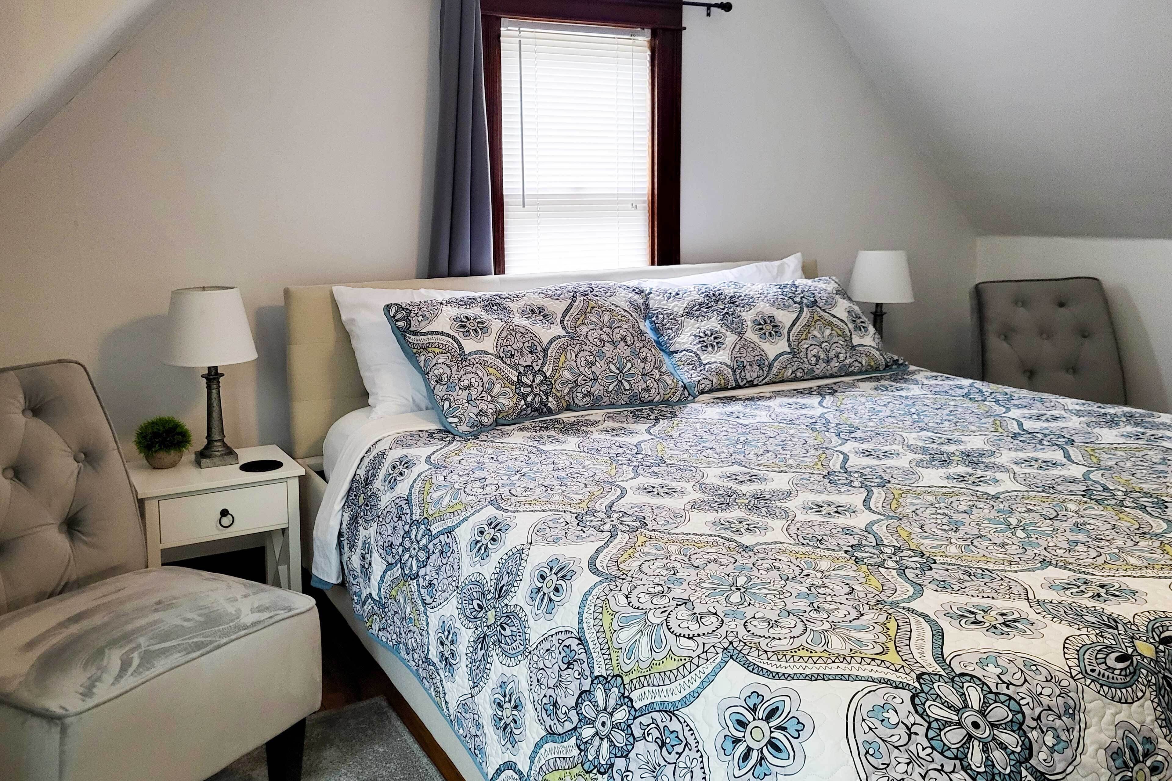 A bedroom with a large bed, a bedside table, a lamp, a chair, and a window with curtains.