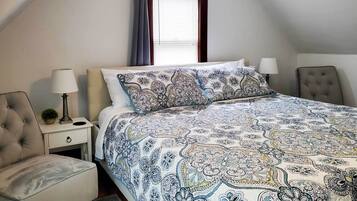 A bedroom with a large bed, a bedside table, a lamp, a chair, and a window with curtains.