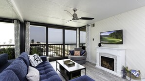 A living room with a large window, a fireplace, a ceiling fan, and a television on the wall. - Sea Watch 702 by Seaside Vacations & Sales (Ocean City)