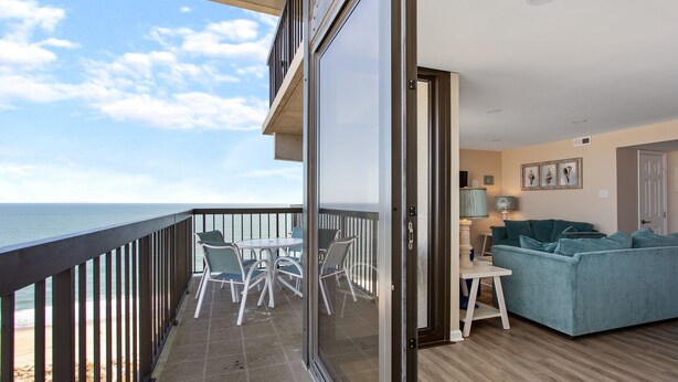 Un balcón con vista al mar, un comedor con sillas y mesa, y una sala de estar con sofá y lámpara.