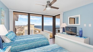 Un dormitorio con vista al océano, un ventilador de techo, un televisor y una cama con ropa de cama azul.