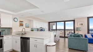 Cocina moderna con armarios blancos, lavavajillas y una isla grande. Cuenta con un comedor con mesa y sillas, y un balcón con vistas.