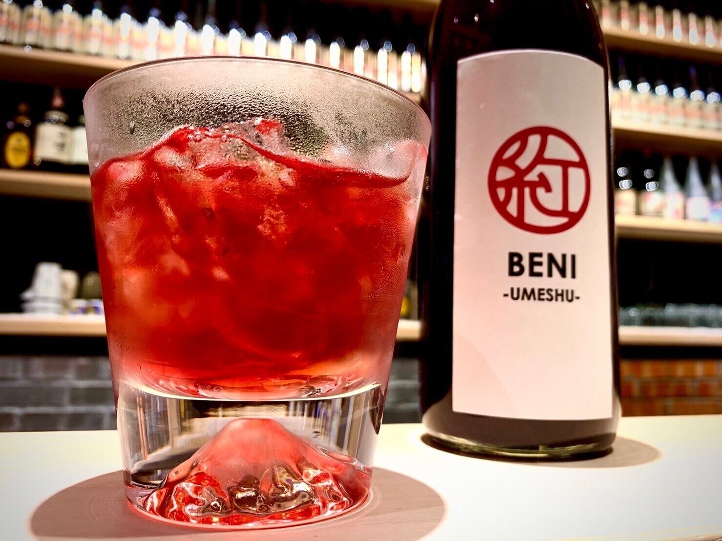 Un vaso de bebida roja con cubitos de hielo, una botella con la etiqueta "BENI -UMESHU-", y un bar de fondo.