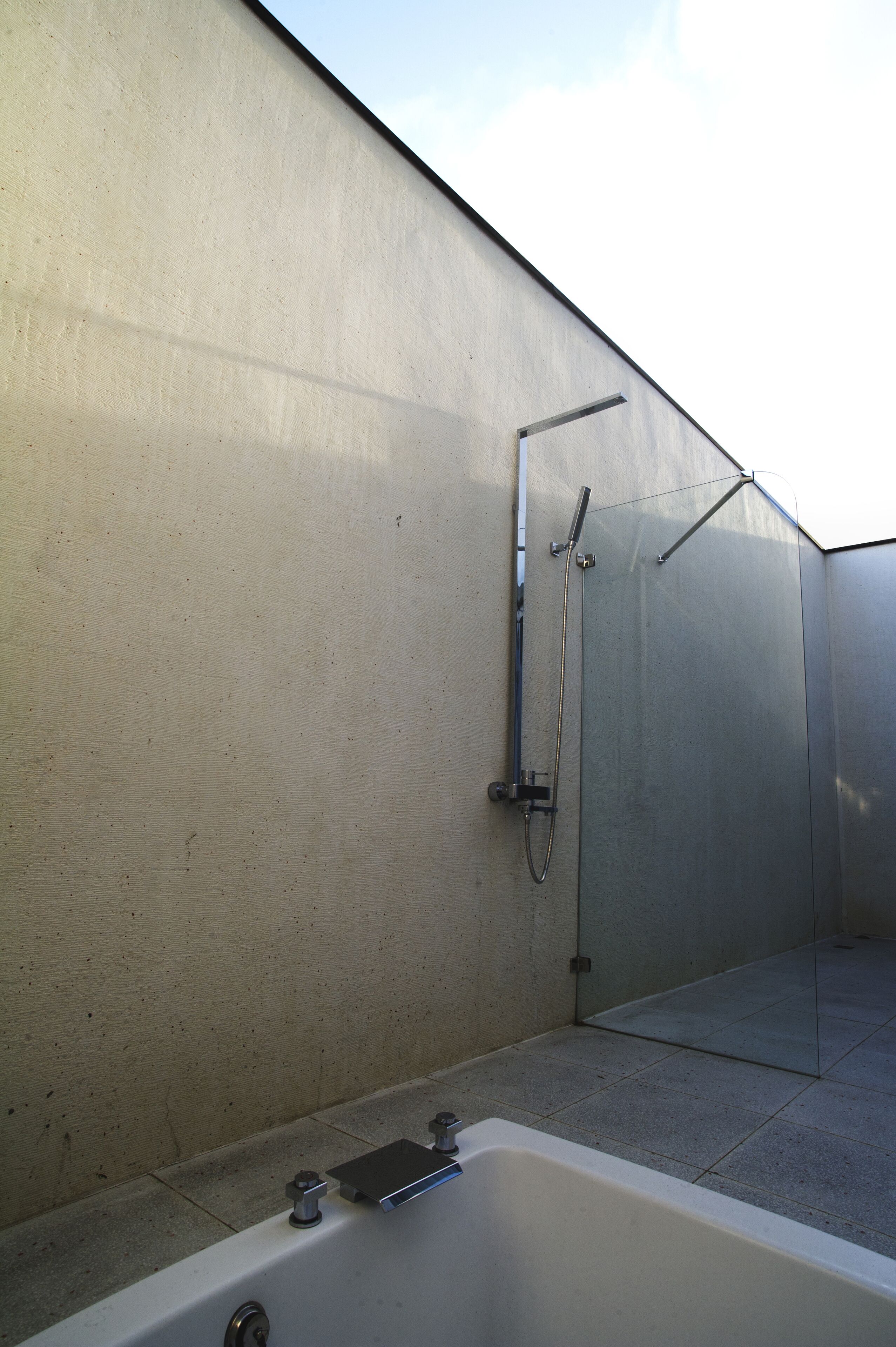 A modern outdoor shower area with a glass enclosure, a white bathtub, and a metal showerhead.