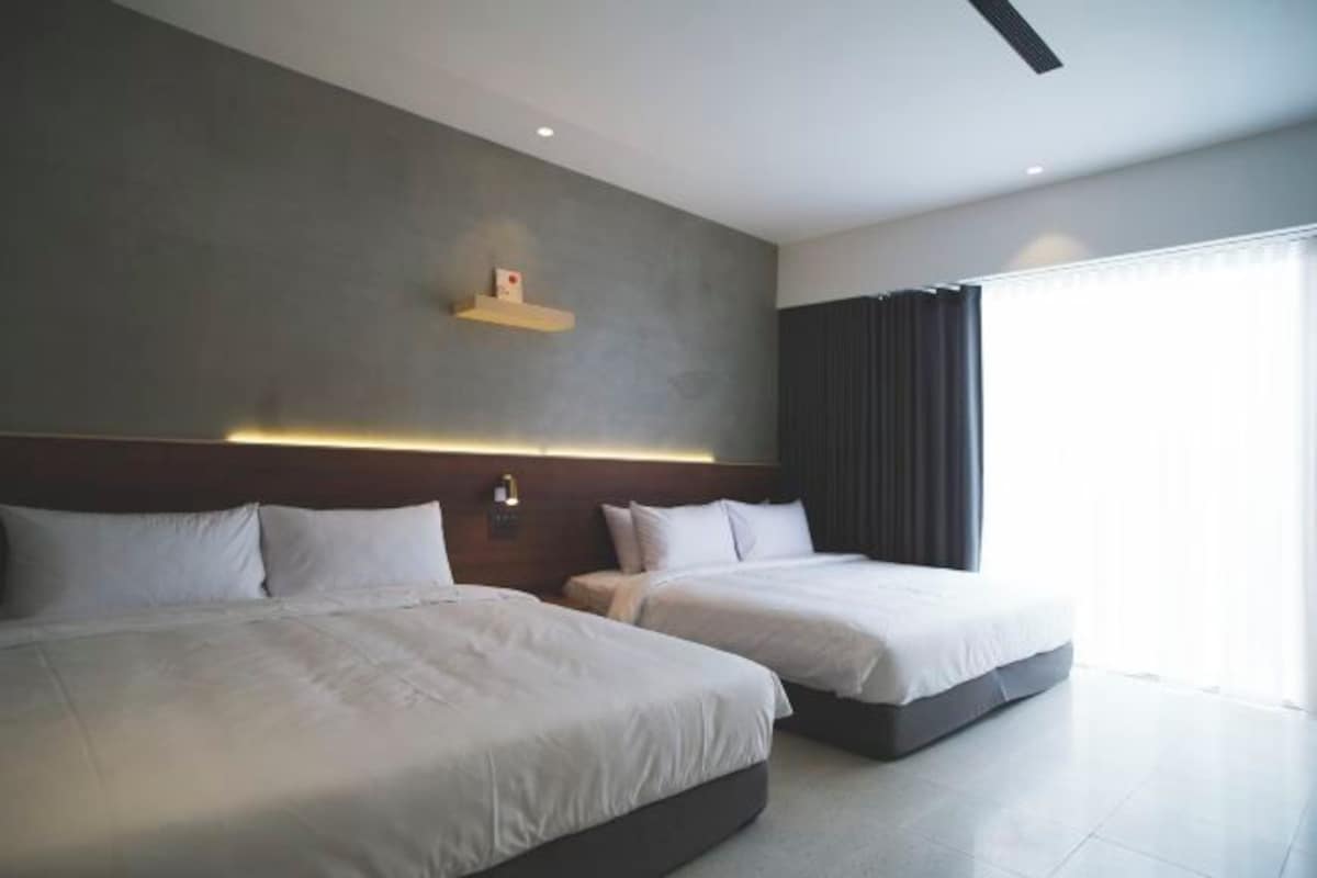 A modern hotel room with two beds, a large window with curtains, and a wall-mounted shelf.
