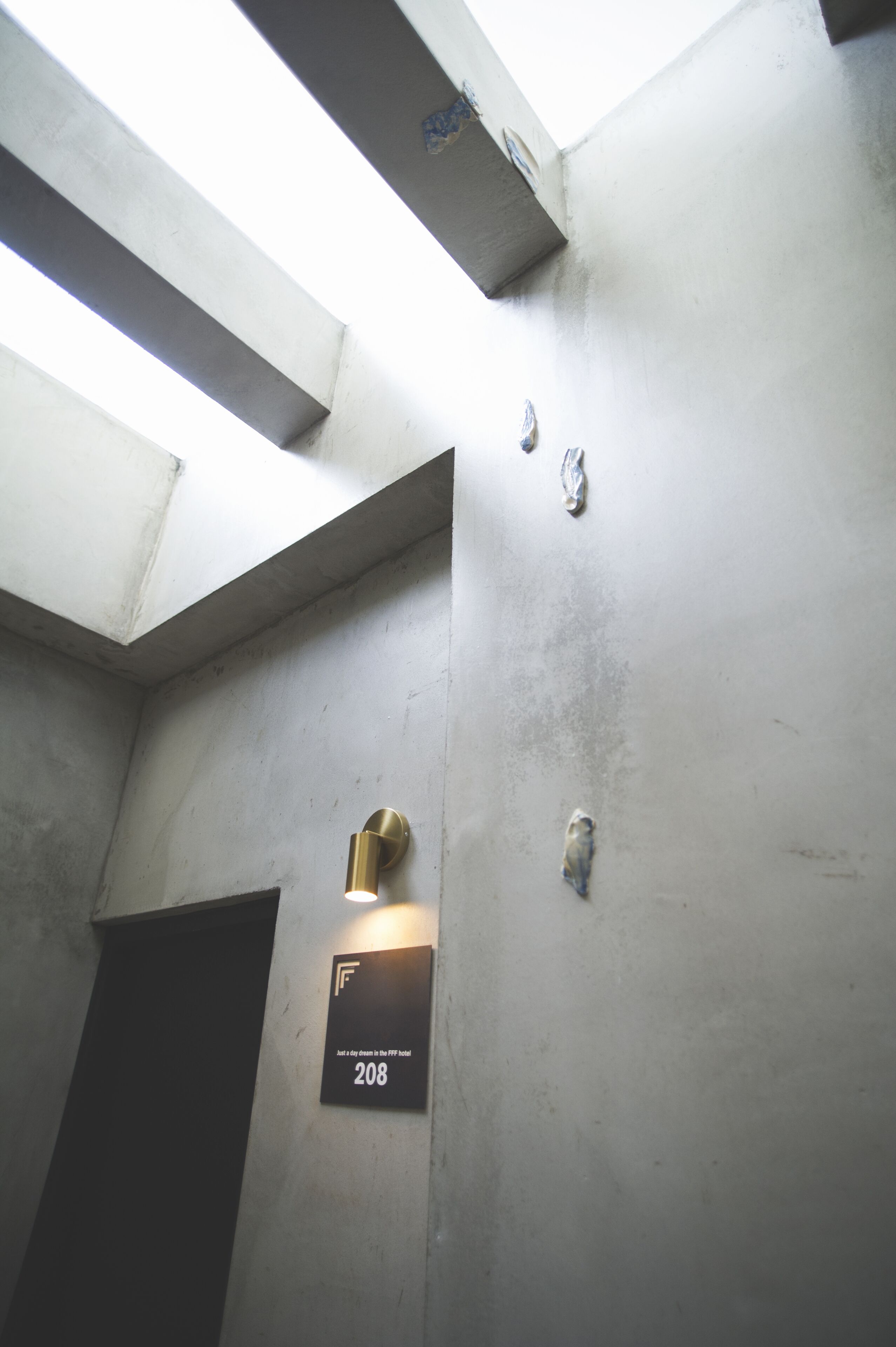 Un corridoio con una parete in cemento, una porta con la scritta '208' e un lampadario.