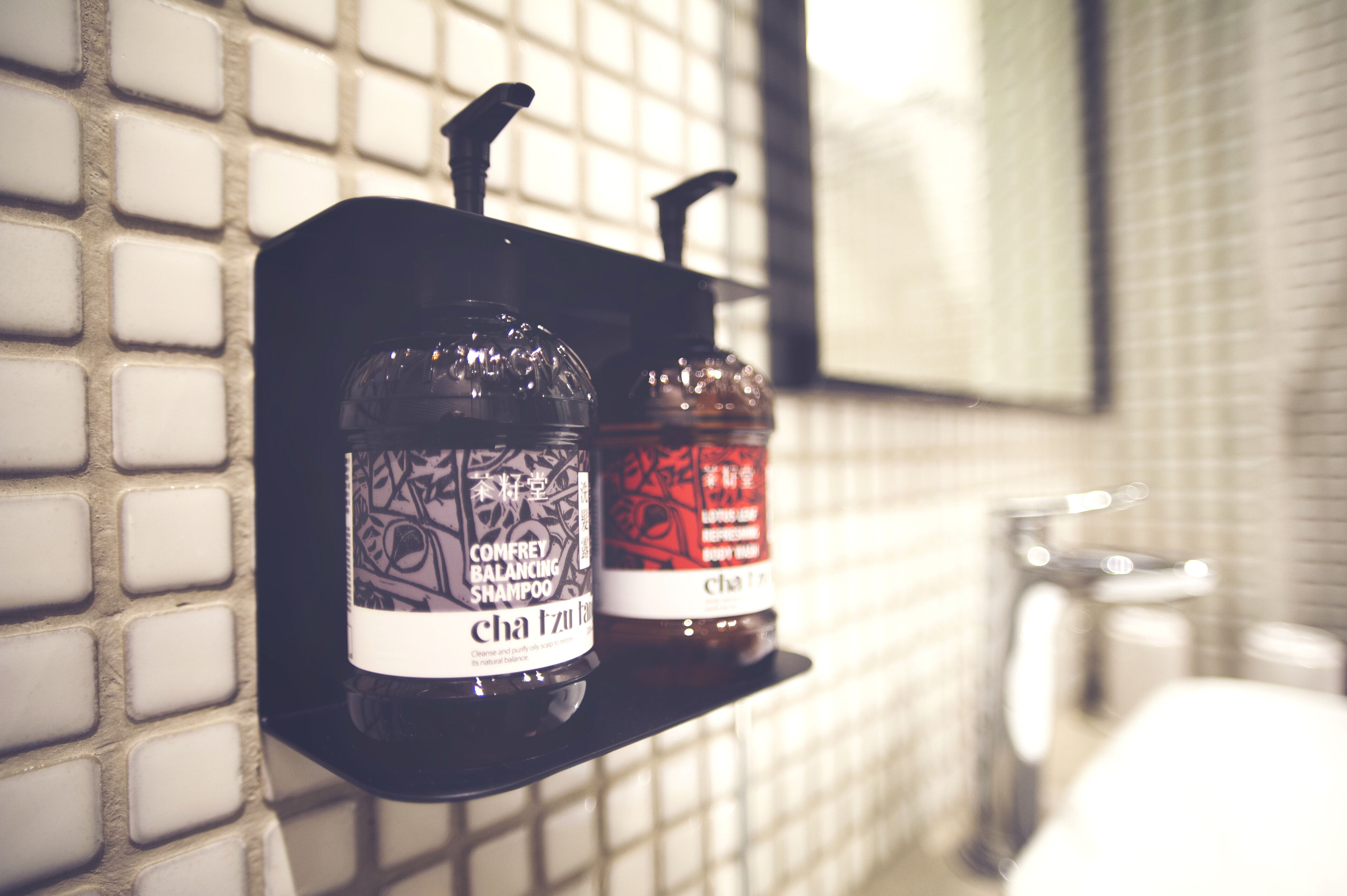 Deux bouteilles de shampoing sur une étagère noire, dans une salle de bain dont un mur est recouvert de carreaux.