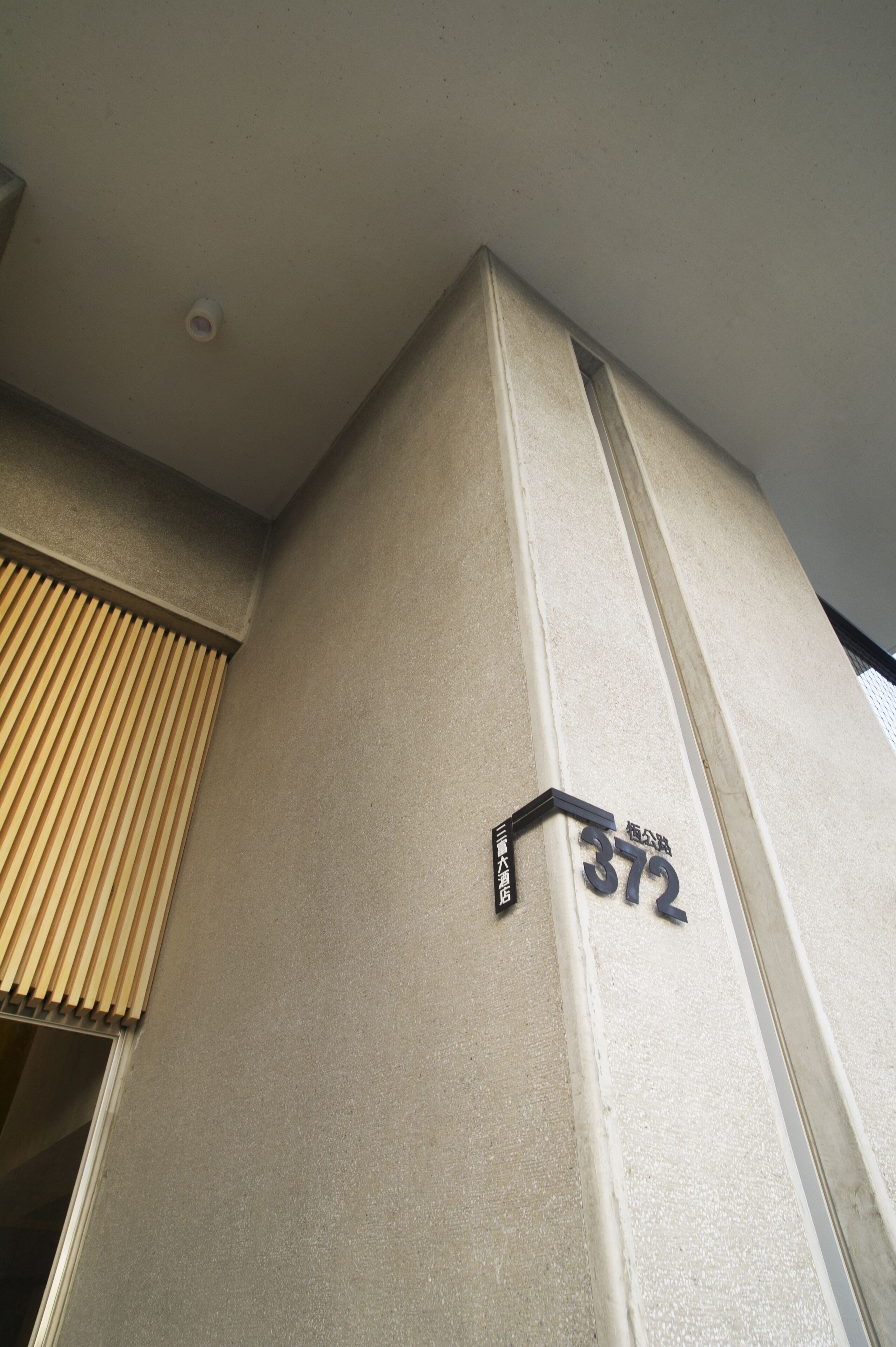 A corner of a building with a beige wall and a window with vertical blinds. A sign with the number "372" is visible.