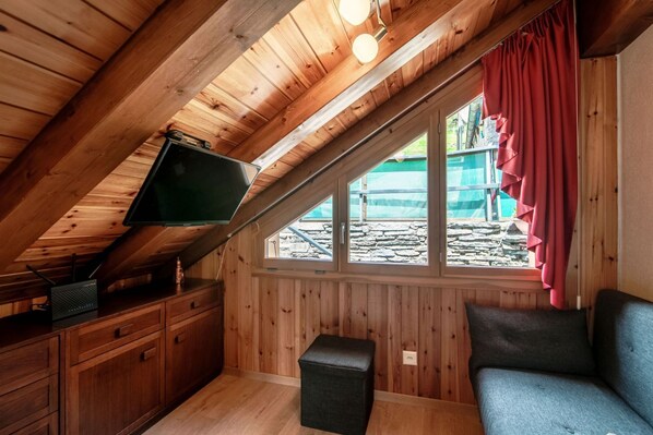 A cozy attic bedroom with wooden walls, a slanted ceiling, a television on a dresser, a window with red curtains, and a small ottoman.