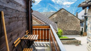 Een houten balkon met een tafel en stoelen, een stenen gebouw op de achtergrond, en een bergachtig landschap.