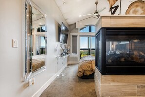 A bedroom with a large mirror, a TV, a fireplace, and a view of the outdoors.