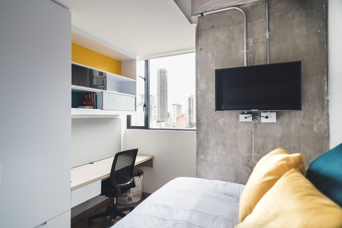 A modern bedroom with a bed, a desk, a chair, a TV mounted on a concrete wall, and a window with a city view.