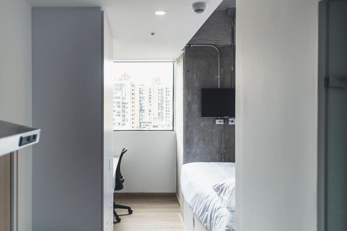 A narrow, modern hotel room with a bed, a desk, and a chair.