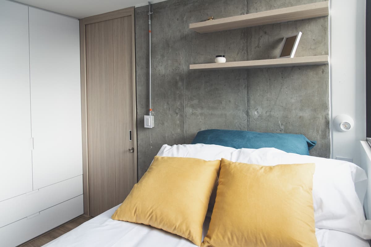 A bedroom with a bed, two pillows, a wooden wardrobe, and a concrete wall with shelves.