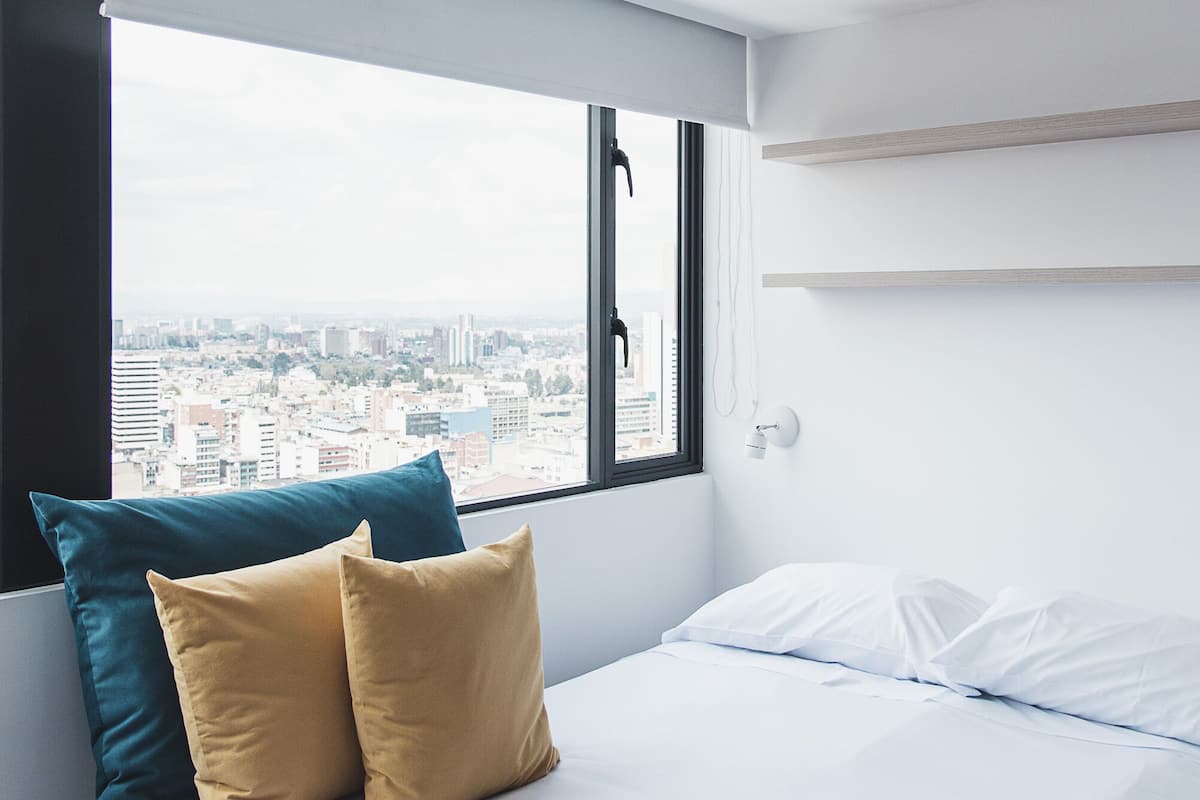 A modern bedroom with a city view, a bed with white and mustard pillows, and a window with a view of buildings.