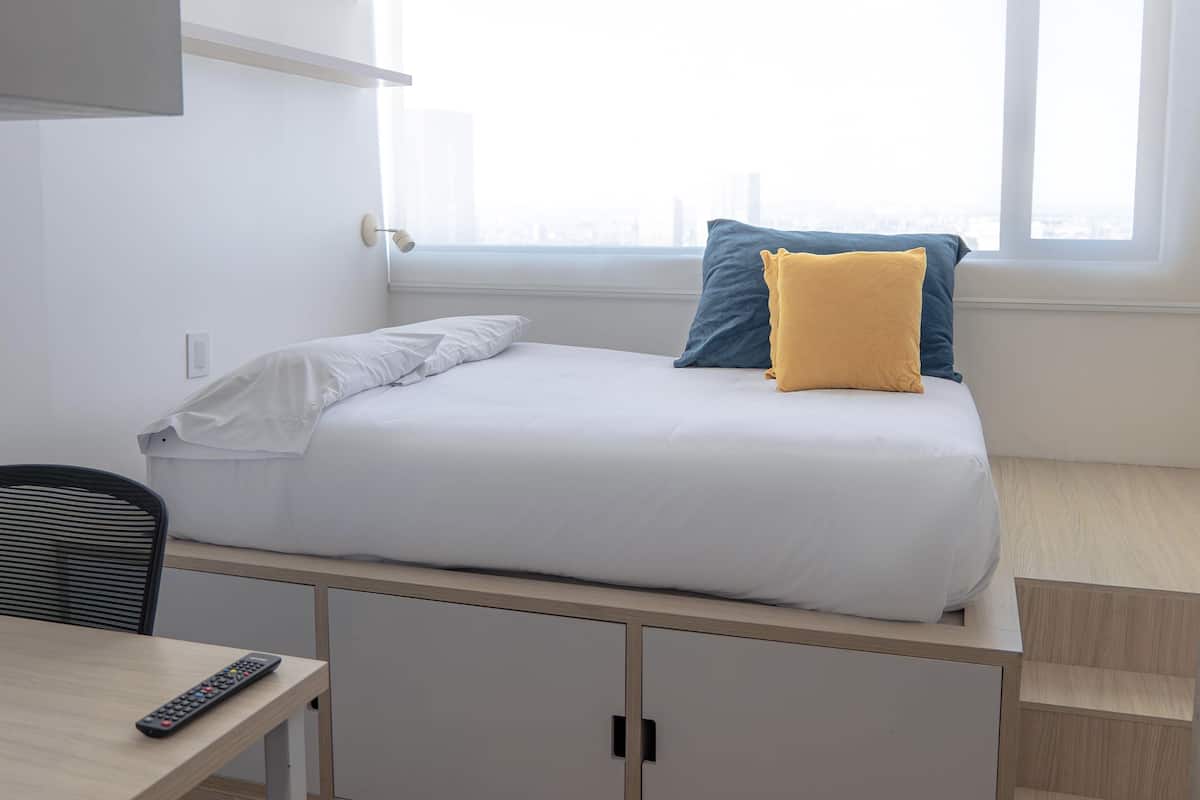 A single bed with white and blue pillows, a small table with a remote control, and a window with a city view.