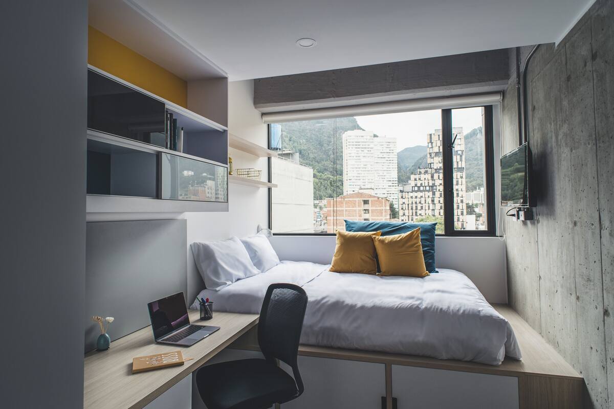 A modern bedroom with a bed, desk, and shelves, featuring a city view through a large window.