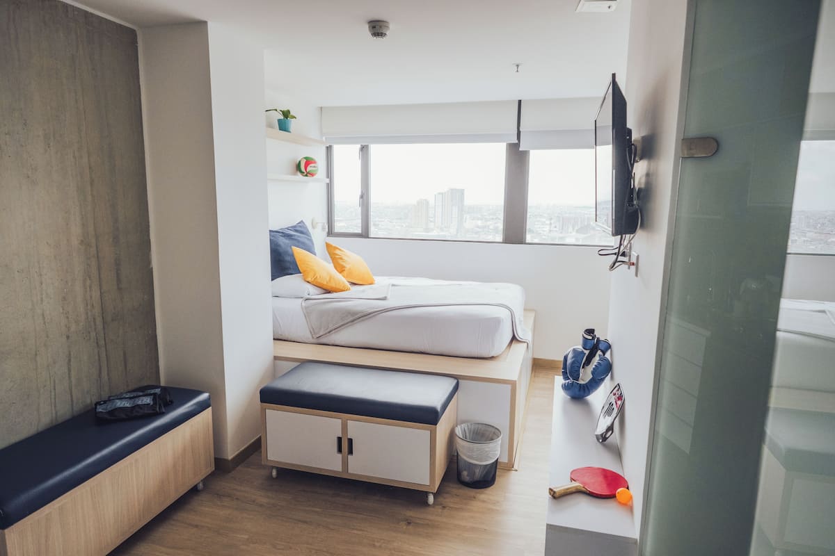 A compact bedroom with a bed, bedside table, and a view of the city through a large window.