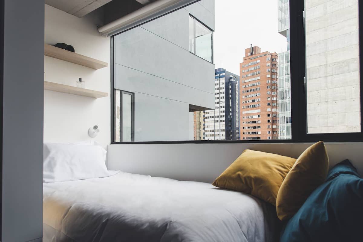 A modern hotel room with a large window, a bed with white and blue bedding, and a wooden shelf with a bottle.