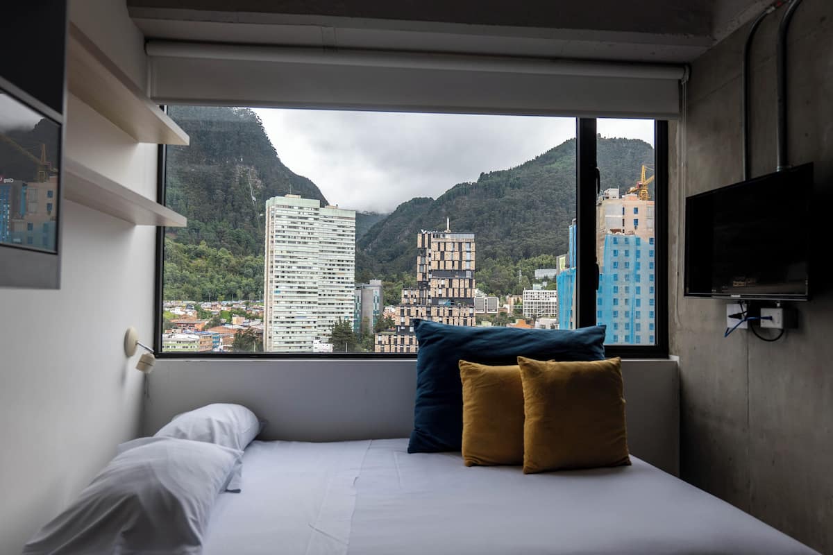 A bedroom with a bed, pillows, a TV, and a window with a cityscape view.