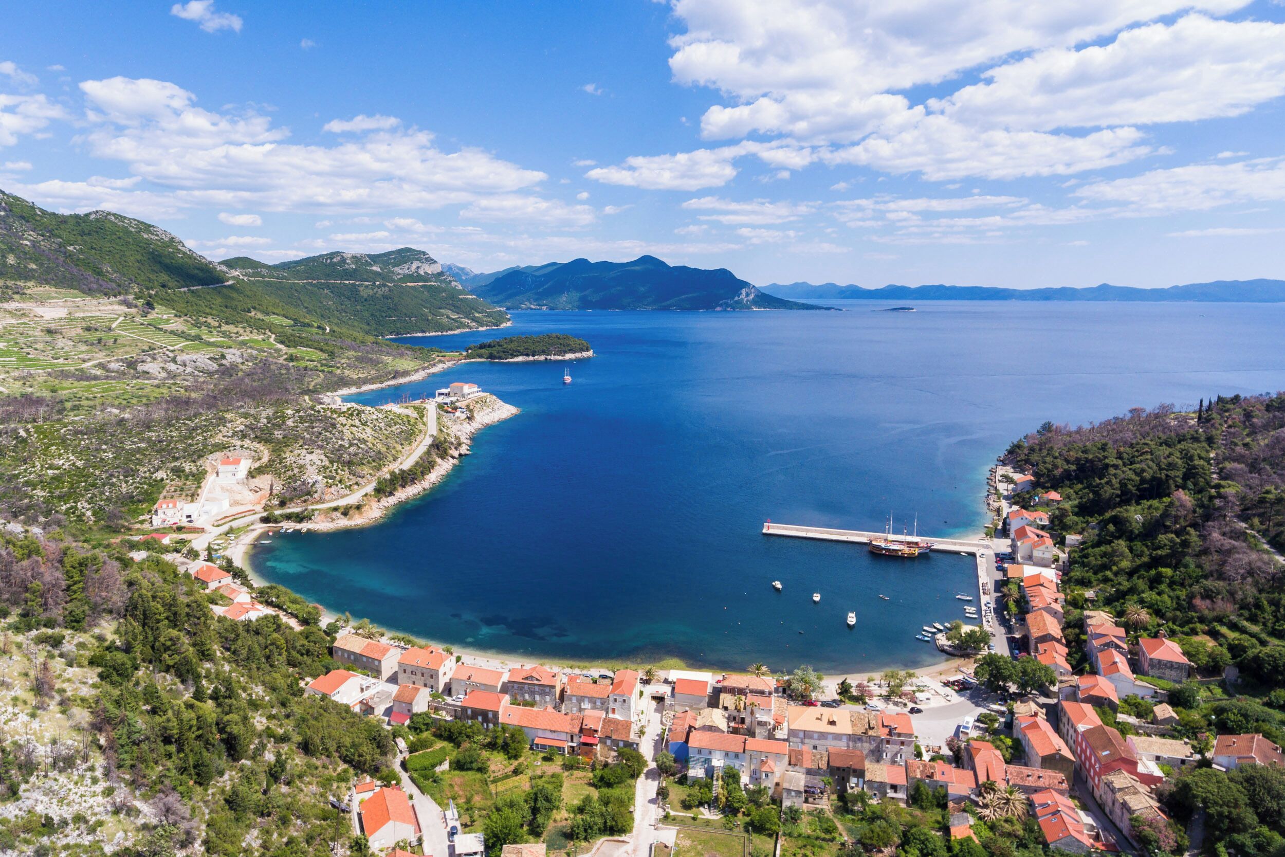 Vista aérea de uma vila costeira com um porto de iates, edifícios com telhados vermelhos e um mar azul límpido.
