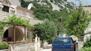 Eine Wohnstraße mit einem blauen Auto am Straßenrand, einer Steinmauer mit Blumentöpfen und einem Berg im Hintergrund.