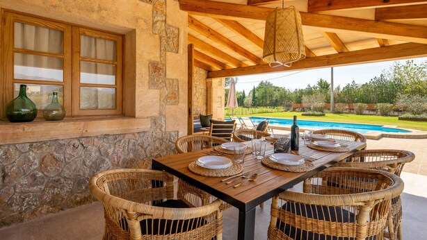 A dining area with a stone wall, wooden ceiling, and wicker chairs. - Villa Can Canavaret by SunVillas Mallorca (Pollença)