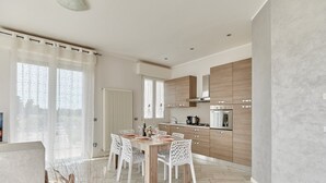A modern kitchen with wooden cabinets, a dining table with chairs, and a large window with sheer curtains. - MINERVA HOLIDAYHOME - MIMOSA APARTMENT (Manerba del Garda)