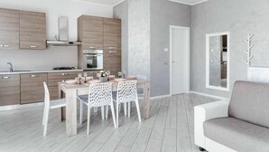 A modern kitchen with wooden cabinets, a dining area with a table and chairs, and a grey sofa in a living room. - MINERVA HOLIDAYHOME - MIMOSA APARTMENT (Manerba del Garda)