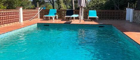 Ein rechteckiger Swimmingpool mit klarem, blauem Wasser, umgeben von einer Holzterrasse. Drei blaue Liegestühle und ein Sonnenschirm befinden sich dort.